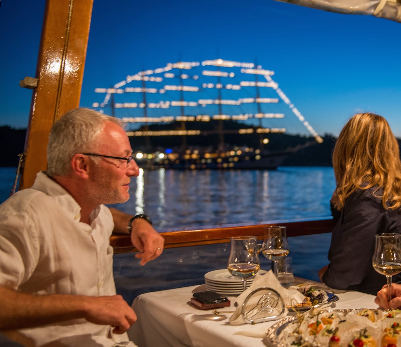 Guests at an evening dinner on a terrace above the sea, with the illuminated Dubrovnik bridge behind